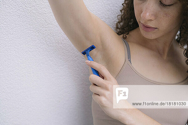 Young woman shaving armpit in front of wall