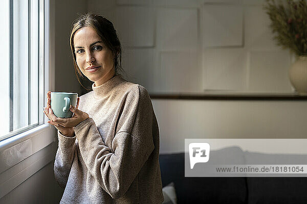 Smiling young woman with cup of tea standing by window at home