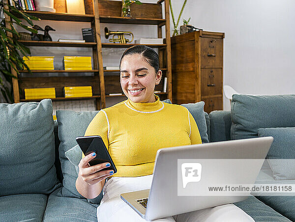 Smiling businesswoman using smart phone sitting with laptop on sofa