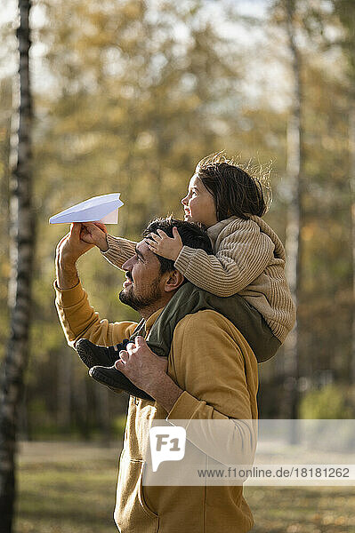 Father carrying boy holding paper airplane on shoulders in park