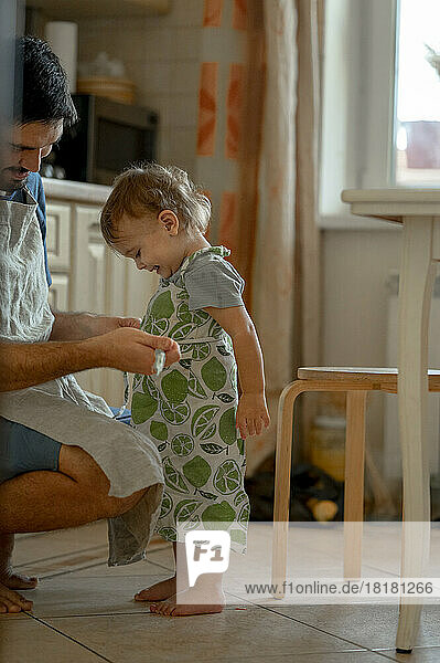 Father tying apron to baby boy in kitchen at home