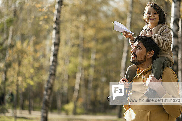 Father carrying son holding paper airplane on shoulders in park