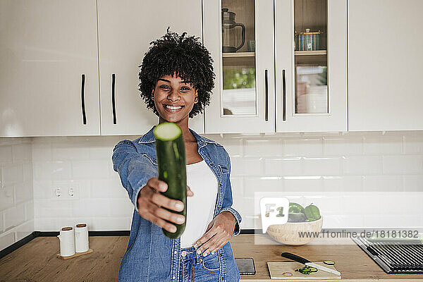 Happy young woman showing zucchini leaning on kitchen counter at home
