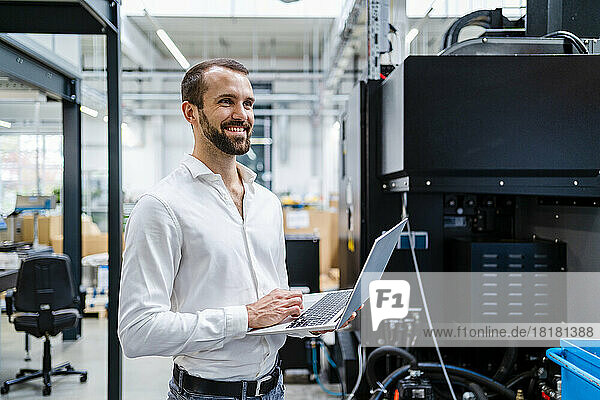 Smiling young businessman with laptop near machinery in factory