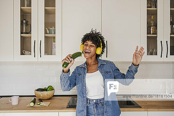 Cheerful woman wearing wireless headphones singing with zucchini in kitchen at home