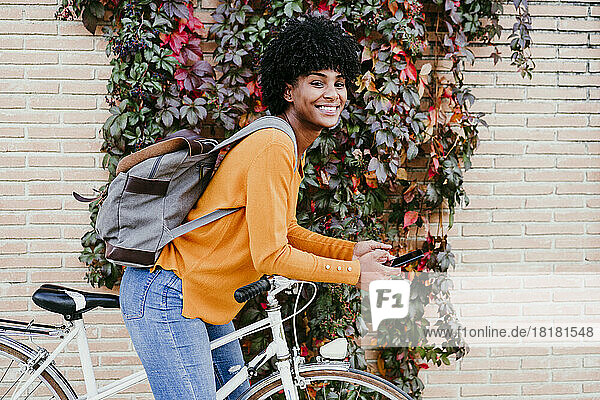 Smiling young woman with backpack holding smart phone on bicycle