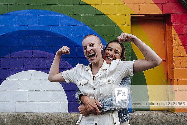 Glückliche lesbische Frau umarmt nicht-binäre Person und lässt vor der Regenbogenwand Muskeln spielen