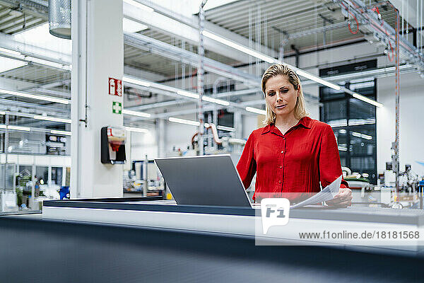 Businesswoman reviewing documents by laptop in factory
