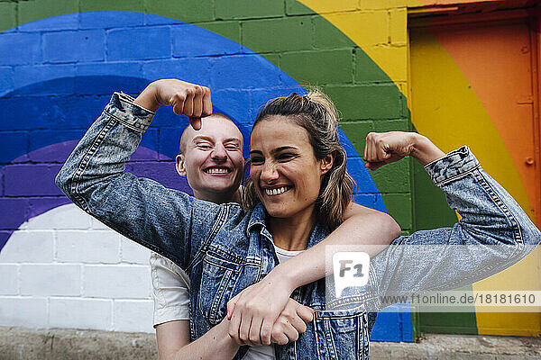 Nicht-binäre Person umarmt eine lesbische Frau  die vor der Regenbogenwand ihre Muskeln spielen lässt