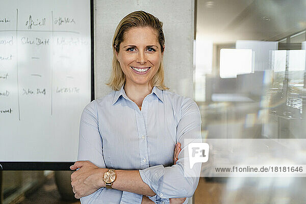 Happy businesswoman with arms crossed leaning on column at workplace