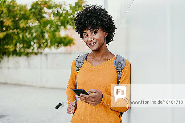 Smiling young woman with mobile phone by wall