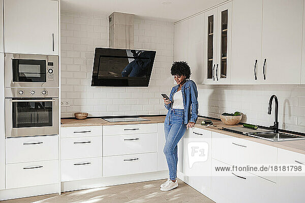 Young woman using smart phone leaning on kitchen counter at home