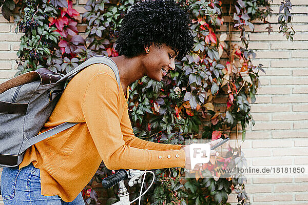 Young woman with backpack using smart phone by ivy wall