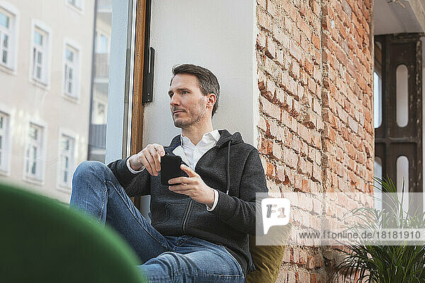 Nachdenklicher Geschäftsmann mit Smartphone sitzt auf der Fensterbank