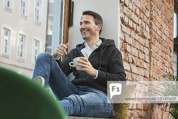 Happy businessman with in-ear headphones holding coffee cup sitting on window sill
