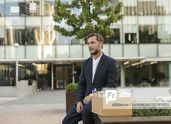 Trauriger Geschäftsmann sitzt mit Karton vor Bürogebäude