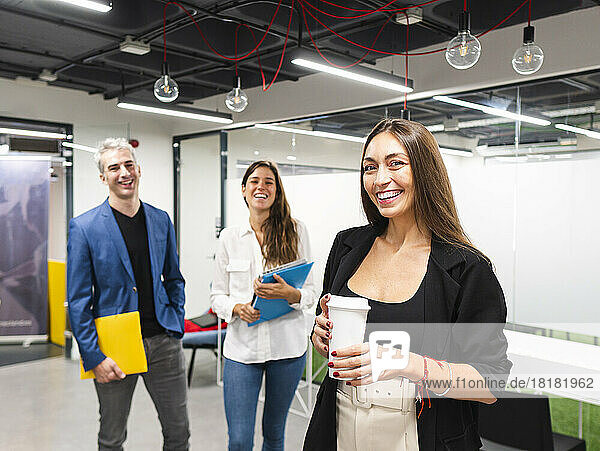 Glückliche Geschäftsfrau hält Einwegkaffeetasse mit Kollegen im Hintergrund