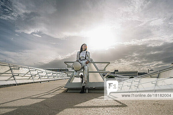 Happy astronaut holding space helmet leaning on railing in front of sky