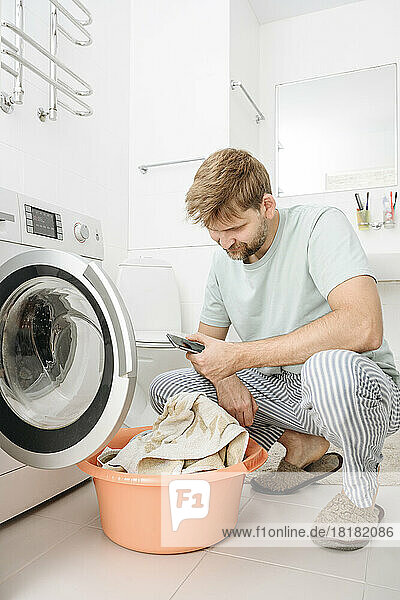 Man using smart phone by washing machine in bathroom at home