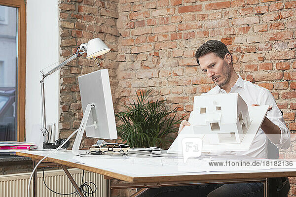 Mature architect examining house model sitting at desk in office