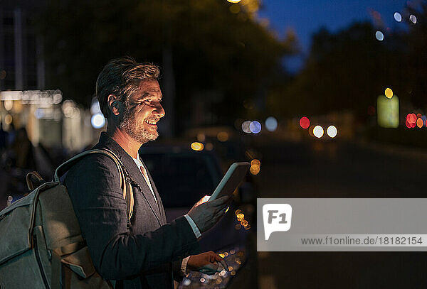 Smiling businessman with backpack holding smart phone at street