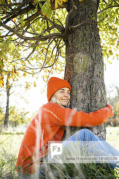 Lächelnde Frau umarmt Baumstamm am Wochenende im Herbst