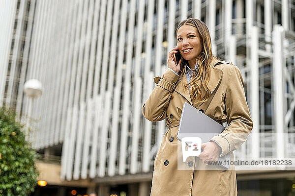 Smiling businesswoman holding laptop talking on mobile phone in front of building