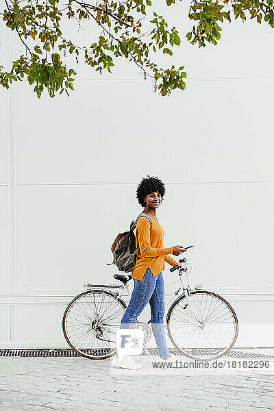 Young woman holding smart phone walking with bicycle by wall