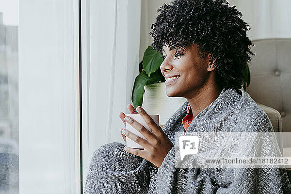 Happy young woman with coffee cup sitting by window at home