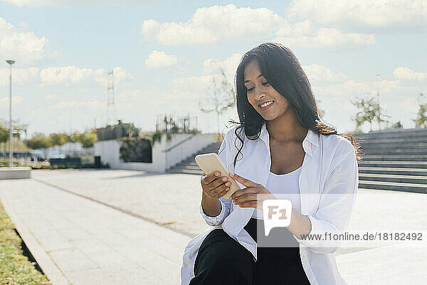 Glückliche junge Frau  die an einem sonnigen Tag im Freien sitzt und ihr Smartphone benutzt