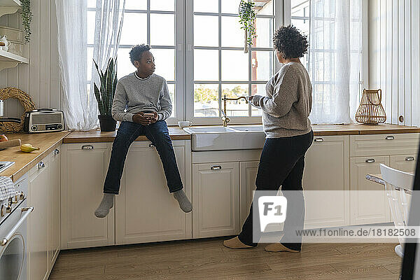 Mutter wäscht Utensilien  während ihr Sohn zu Hause auf der Küchentheke neben der Spüle sitzt