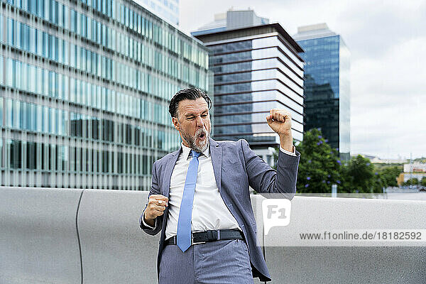 Happy mature businessman gesturing fists standing in front of building