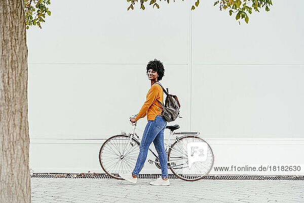 Smiling woman with backpack wheeling bicycle on footpath