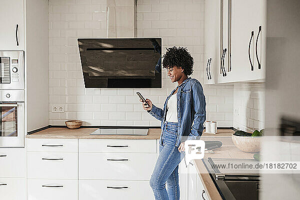 Happy woman using smart phone leaning on kitchen counter at home