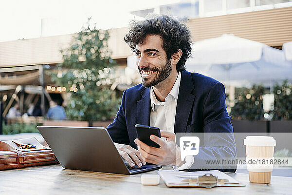 Smiling businessman with smart phone and laptop working from cafe