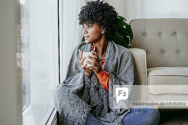 Junge Frau mit Kaffeetasse sitzt zu Hause am Fenster