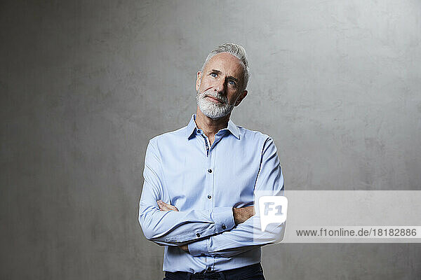 Contemplative mature man with arms crossed standing in front of grey background