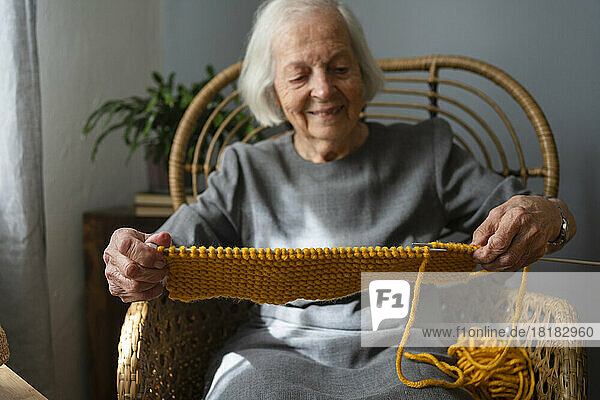 Smiling senior woman knitting scarf on chair at home
