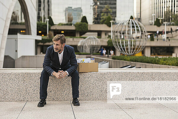 Trauriger Geschäftsmann mit Karton sitzt an der Wand im Büropark