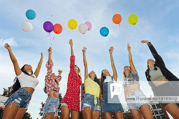 Friends with hands raised holding colorful balloons under blue sky