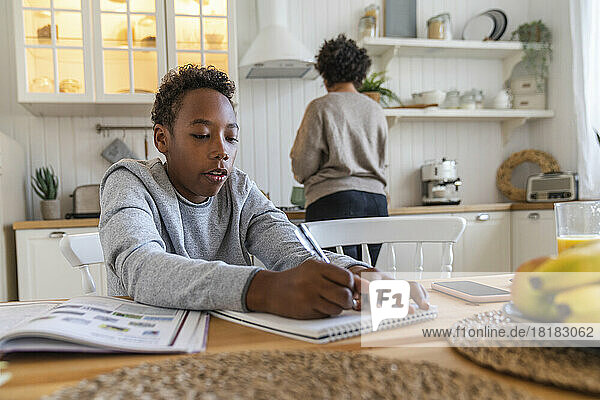 Boy writing on book with mother in background working at home