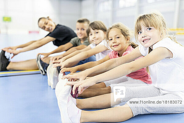 Smiling elementary students doing exercise sitting side by side with teacher at school sports court