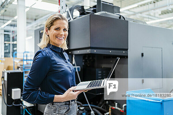 Smiling businesswoman with laptop standing by machinery in factory