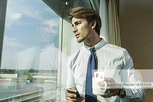 Portrait of young businessman with smartphone and mug looking through window