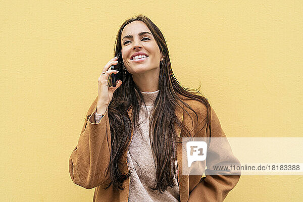 Happy young woman talking on mobile phone in front of yellow wall