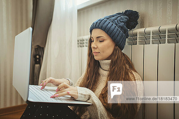Woman wearing knit hat using laptop leaning on radiator at home