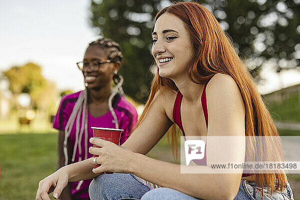 Happy redhead woman sitting with drink by friend at park