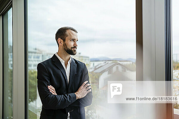 Young businessman with arms crossed looking out of window at office