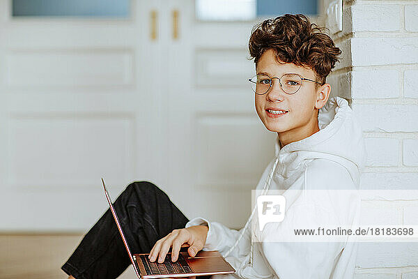 Smiling boy with laptop sitting by wall