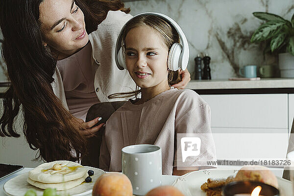 Smiling woman looking at daughter listening to music through wireless headphones